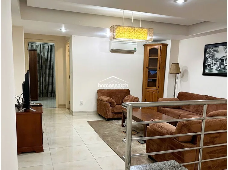 The living room is furnished with a brown fabric sofa set, a wooden coffee table, and a matching corner display cabinet. A wall-mounted air conditioning unit is present, and the area flows directly into a hallway that leads to the bedroom and other parts of the property.