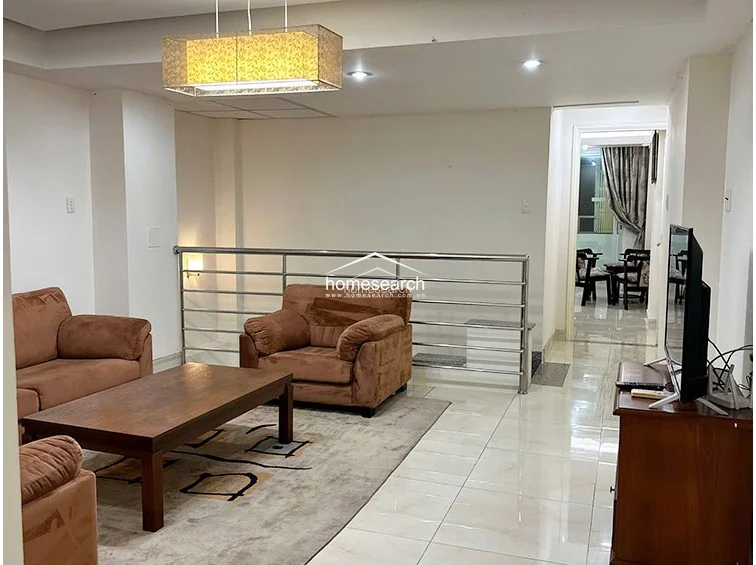 This living area features a split-level layout with a stainless steel railing separating the lounge from the circulation path. The space is finished with glossy light-colored floor tiles, white walls, and a prominent rectangular pendant light fixture, creating a clean, functional aesthetic.