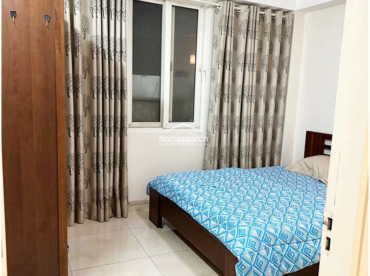 This compact bedroom features a double bed with a blue geometric-patterned quilt and a wooden headboard. Natural light enters through a window dressed in floor-to-ceiling curtains with a tree-motif pattern, and the room includes a wooden wardrobe for storage.