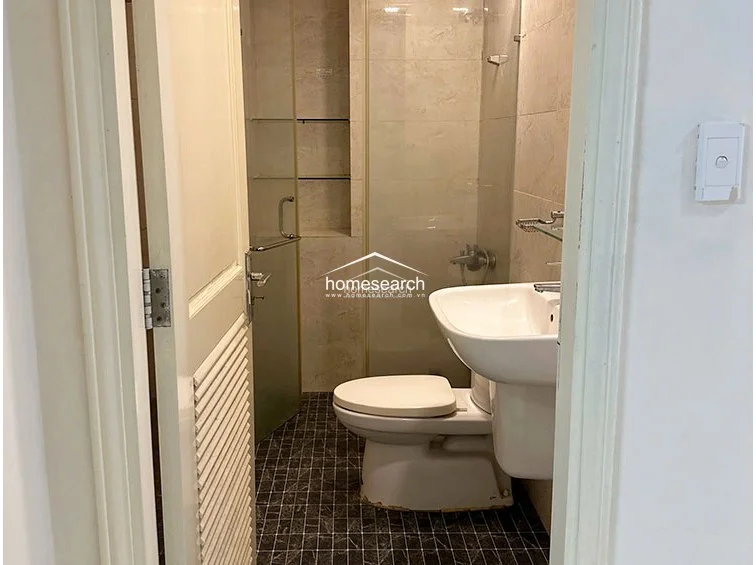 This bathroom features a compact, functional layout with a pedestal sink, a white toilet, and a glass-enclosed shower area with built-in shelving. The space is finished with contrasting dark-patterned floor tiles and light-toned wall tiles, showing a well-maintained, utilitarian condition.