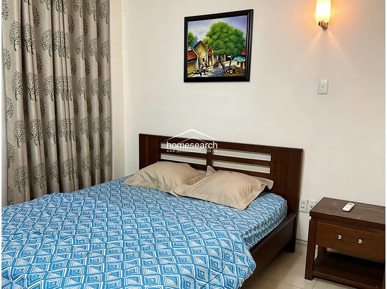 This bedroom view highlights a wooden bed frame, a matching nightstand, and a wall-mounted reading lamp. The room is finished with white walls and a piece of framed art, maintaining a consistent, neutral, and well-maintained style throughout the sleeping quarters.