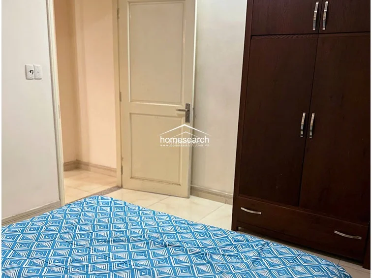 This bedroom features a large, dark wood wardrobe providing ample storage and a bed with a vibrant blue geometric-patterned cover. The room is finished with neutral-toned walls and light-colored tile flooring, creating a clean, functional space that connects directly to the hallway.
