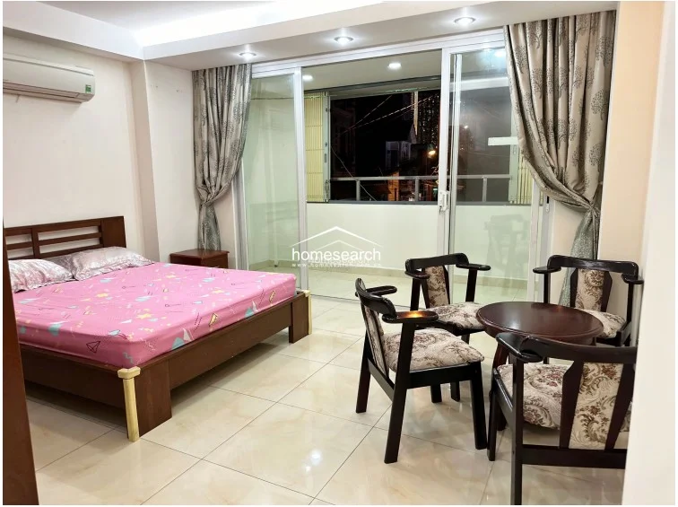 This bedroom features a large sliding glass door that provides access to a private balcony and allows for significant natural light. The room is furnished with a wooden bed frame, a small circular seating area, and an AC unit, all set against a neutral backdrop with patterned curtains.