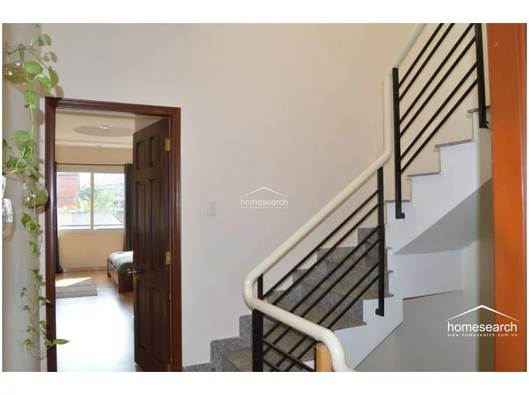 This image shows a staircase landing area featuring light-colored walls and a modern staircase with a white frame and black metal railings. The flooring appears to be a speckled stone or tile, and the open doorway provides a direct line of sight into a well-lit bedroom with wood-look flooring.