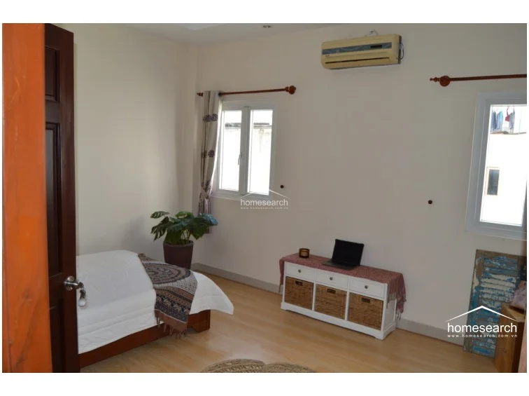 This bedroom is furnished with a single bed, a white storage console with wicker baskets, and a wall-mounted air conditioning unit. The room features light wood-tone flooring and two windows that provide natural light, creating a functional and airy living space.