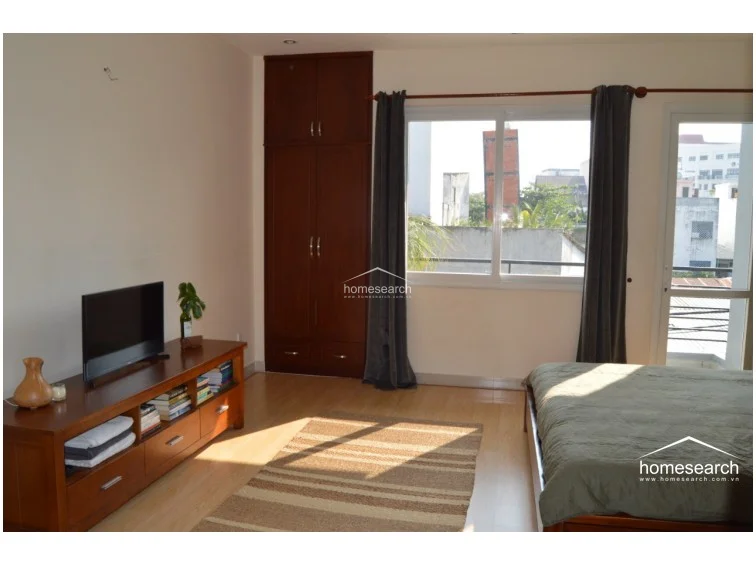This bedroom layout includes a direct connection to a private balcony through a glass door, allowing for significant natural light and urban views. The space is well-organized with a built-in dark wood wardrobe and a wooden media console, complemented by a simple, neutral color palette and light flooring.