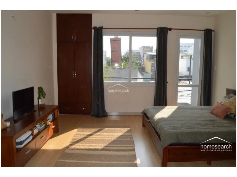 This bedroom features a queen-sized bed, a wooden media console, and a tall built-in dark wood wardrobe for storage. Large windows and a glass door provide ample natural light and access to an outdoor balcony, while the neutral walls and light wood-tone flooring create a clean, minimalist aesthetic.