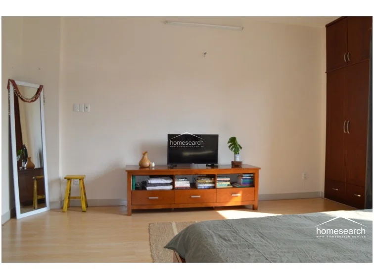 This bedroom features light wood-look flooring and neutral off-white walls, creating a clean and minimalist aesthetic. The room is furnished with a dark wood media console, a matching built-in wardrobe, and a floor-length mirror, all maintained in good condition.