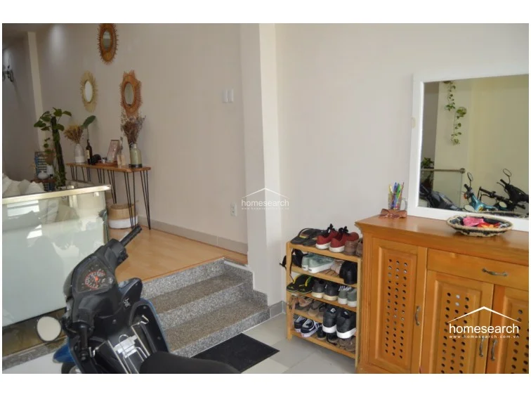The entryway serves as a functional transition space, featuring a stone-stepped elevation that separates the main living area from the door. It includes a wooden shoe cabinet with a large mirror and a small open shelving unit, maintaining a tidy and organized appearance.