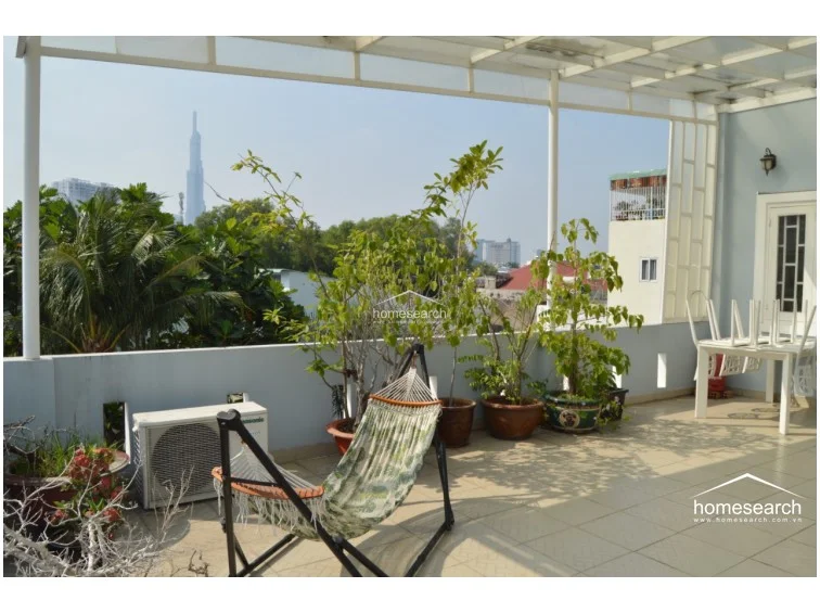 This rooftop terrace continues the open-air theme, featuring a semi-covered structure with a translucent roof that provides protection while maintaining natural light. The area is well-furnished with potted greenery, a hammock, and a dining table, offering a clear, elevated view of the surrounding cityscape.
