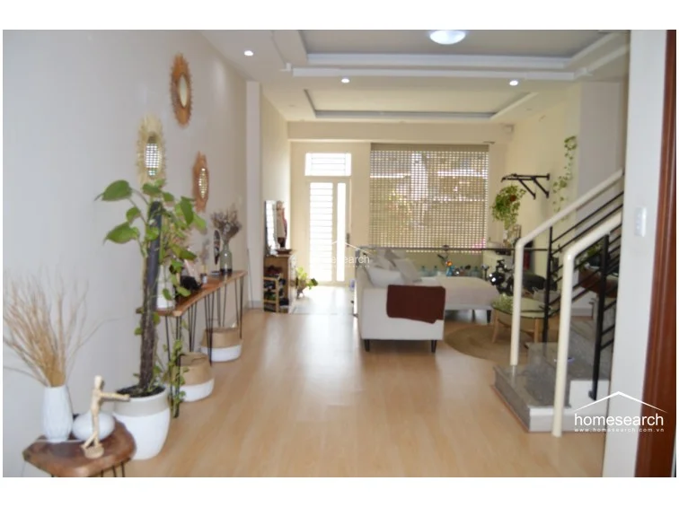 This open-concept living area features light wood-look flooring and a neutral color palette, creating a bright and airy atmosphere. The space flows seamlessly from the entrance toward a living room with a white sofa, illuminated by natural light from a large window with a security roller shutter.