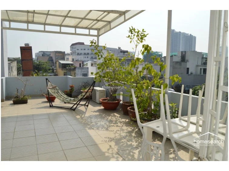 This rooftop space provides a panoramic view of the neighborhood, including high-rise buildings in the distance, and is accessorized with a hammock and several large potted plants. The area is partially covered by a white pergola-style frame, offering a versatile outdoor living space that is well-maintained and bright.