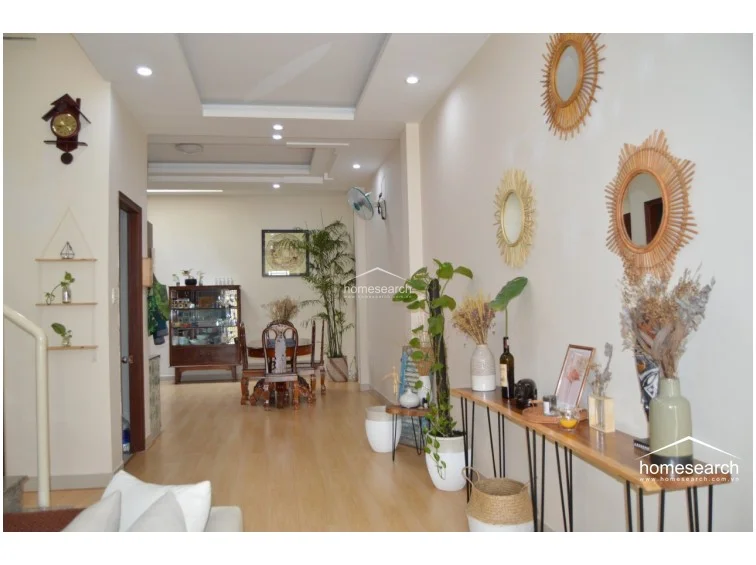The dining and entryway area is decorated with bohemian-style accents, including rattan-framed mirrors and a slim wooden console table. A wooden dining set is positioned in the background, while the space is accented by lush indoor greenery and warm, recessed ceiling lighting.