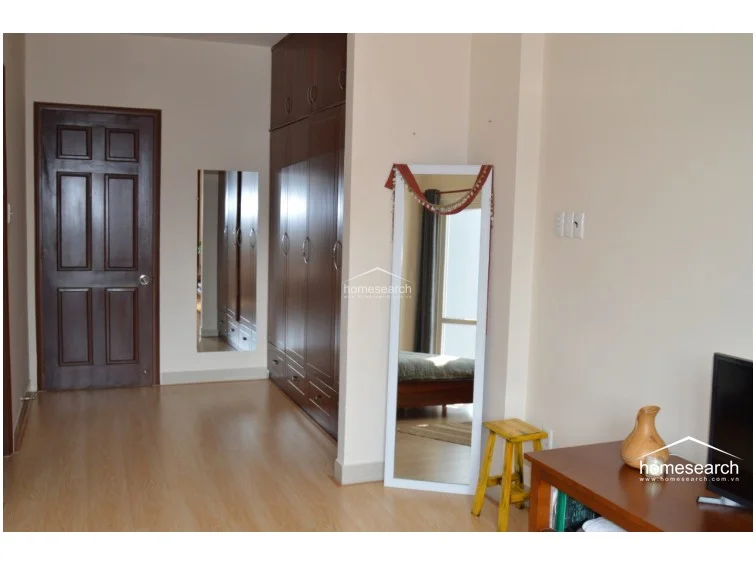 This view highlights a corner of the bedroom featuring a tall, dark wood wardrobe and a full-length floor mirror with a white frame. The space is finished with light wood-tone flooring and cream-colored walls, maintaining a consistent, simple design throughout the room.