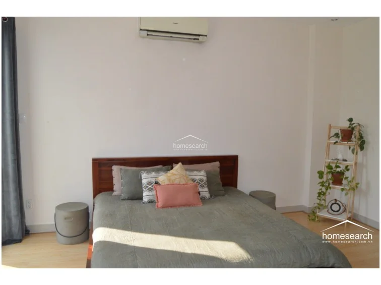 This bedroom setup includes a bed with a dark wood frame, a wall-mounted air conditioning unit, and a decorative wooden ladder shelf for plants. The room is brightly lit by natural light from the adjacent window and features a neutral color palette that emphasizes a calm, uncluttered atmosphere.