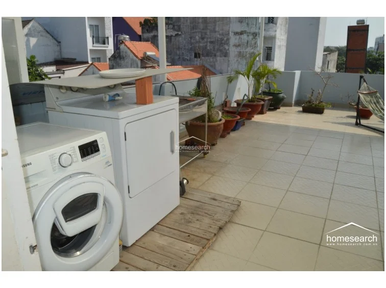 This outdoor rooftop terrace features light-colored floor tiles and serves as a functional laundry area, currently housing a Samsung washing machine and a dryer. The space is decorated with various potted plants and includes a hammock, creating a relaxed, multi-purpose environment with an urban view.