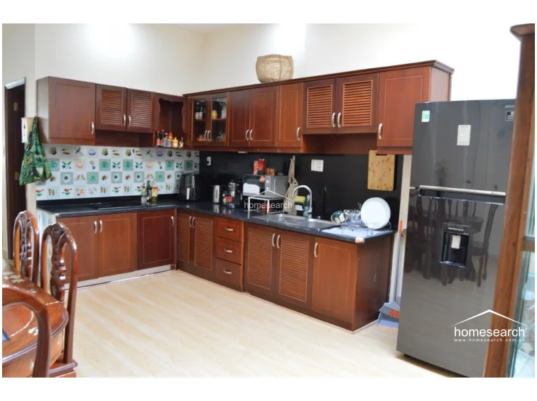 The kitchen features a practical L-shaped layout with dark wood cabinetry and contrasting black countertops. It is well-equipped with a modern black refrigerator, a tiled backsplash with decorative motifs, and ample storage space, all set against light-toned flooring.