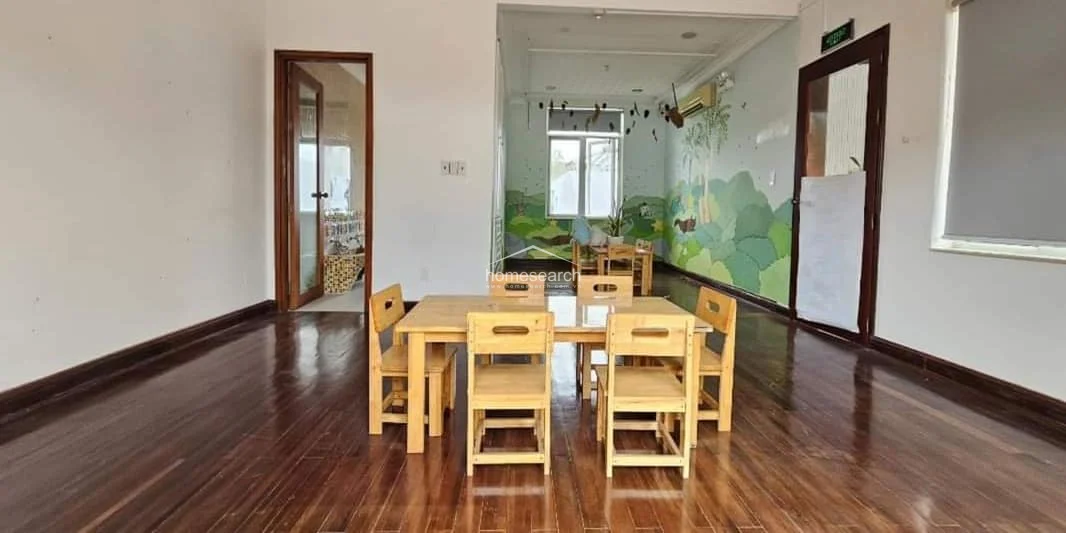 This interior room, currently utilized as a play or learning space, features rich, polished dark wood flooring and high ceilings that enhance the sense of openness. The walls are decorated with playful, nature-themed murals, and the room is equipped with a wall-mounted AC unit, connecting to other areas of the home through wooden-framed glass doors.