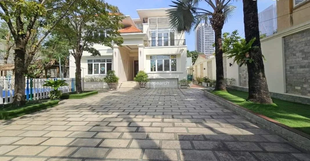 This image showcases the exterior of a modern, multi-story villa featuring a clean, cream-colored facade and large glass windows that suggest bright, airy interiors. The property is approached via a spacious, stone-paved driveway flanked by mature trees and manicured green borders, creating an inviting and well-maintained curb appeal.