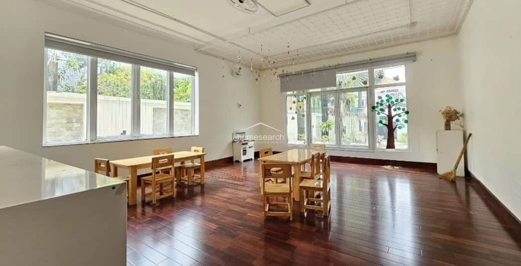 This spacious, light-filled room features rich dark wood flooring and large windows on two walls that provide ample natural light and views of the exterior. Currently utilized as a classroom or play area, the space is finished with simple white walls and a functional ceiling grid, offering a clean, well-maintained environment.