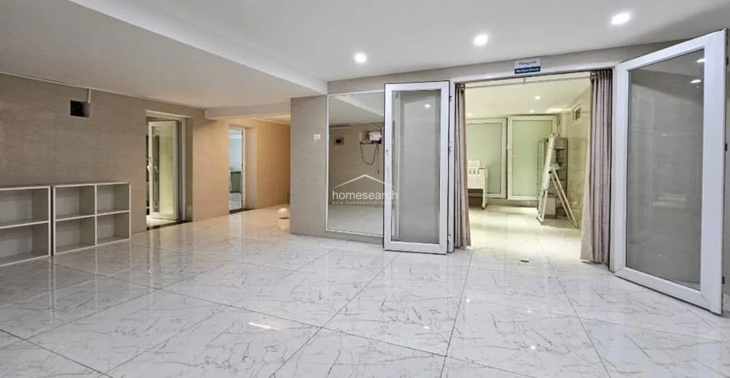 This interior space features polished white marble-look tile flooring and neutral-toned walls, creating a bright and clinical atmosphere. The layout includes large glass-panel doors that open into an adjacent room, with built-in open shelving providing convenient storage and recessed ceiling lighting throughout.