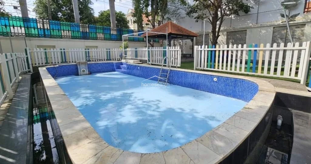 This outdoor swimming pool area is enclosed by a decorative white picket fence and features a tiled pool basin with a built-in ladder for access. A small, tiled gazebo with a terracotta roof sits in the corner, offering a shaded spot for relaxation, though the pool appears to require some cleaning and maintenance.
