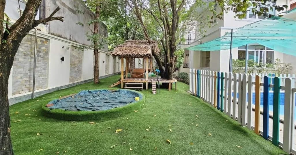 The backyard area is designed as a family-friendly recreational space, featuring a synthetic grass lawn, a small circular play area, and a rustic wooden gazebo nestled under a large tree. A colorful picket fence separates the lawn from the adjacent swimming pool, providing a safe and charming outdoor environment for children.