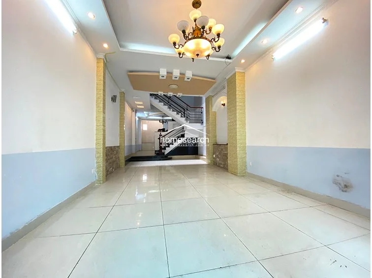 This interior view captures an open-plan living area with polished light-colored floor tiles and a neutral wall color scheme. The space features a prominent central staircase with a dark metal railing and is illuminated by a decorative chandelier and recessed ceiling lighting.