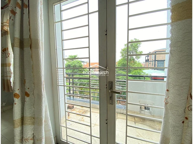 This image shows a balcony access point featuring a white-framed glass door protected by a heavy-duty metal security grille. The balcony provides an outdoor space with views of neighboring residential rooftops and mature trees, offering a sense of openness.
