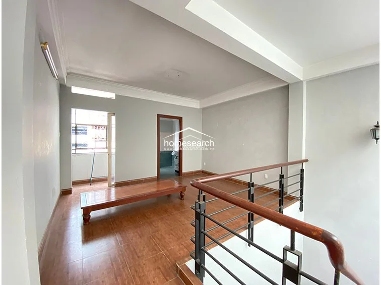 This versatile landing or mezzanine area connects to a balcony and features a simple wooden platform bed frame, suggesting it could function as a lounge or sleeping space. The area maintains a consistent aesthetic with the rest of the home, utilizing neutral wall tones and tile flooring to create a bright, airy flow.