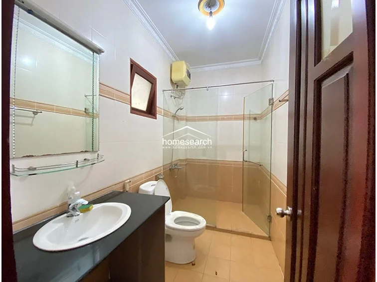 The bathroom is equipped with a glass-enclosed shower stall, a pedestal sink with a dark stone countertop, and a wall-mounted water heater. The space is finished with two-tone beige and white wall tiles and light-colored floor tiles, presenting a functional and well-maintained sanitary area.