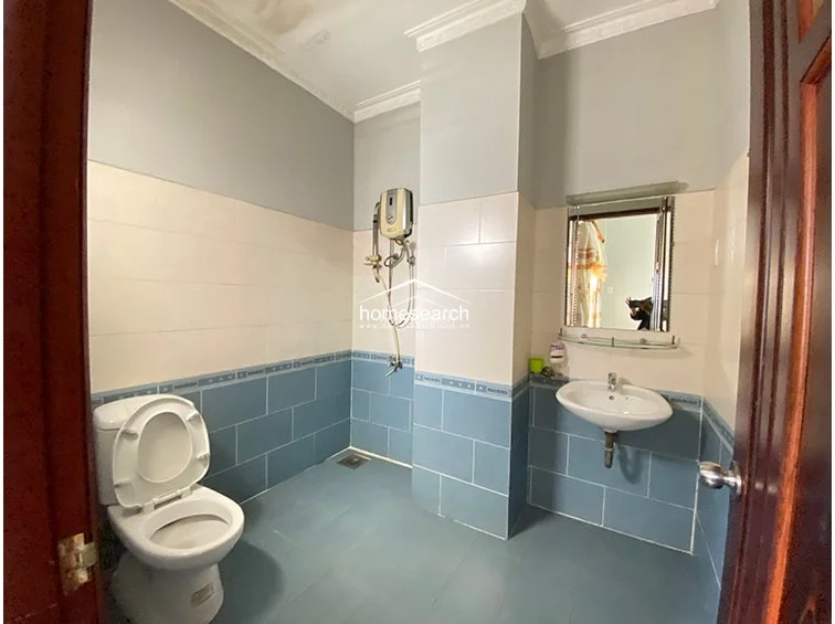 This bathroom features a functional layout with a wet-room style shower area and a wall-mounted sink, finished in a two-tone blue and cream tile scheme. The space is maintained in fair condition and includes a wall-mounted electric water heater, a simple mirror, and a standard white ceramic toilet.
