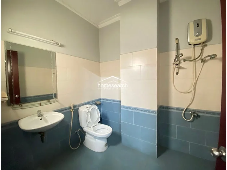 The bathroom maintains a consistent design aesthetic with blue floor and lower-wall tiles contrasted by cream upper tiles and light grey paint. It is equipped with a wall-mounted shower unit, a pedestal sink, and a large mirror, providing a basic and utilitarian space with standard fixtures.