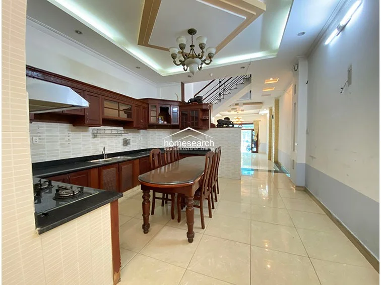 This image displays a spacious kitchen and dining area integrated into the open floor plan, featuring traditional dark wood cabinetry and a matching dining set. The kitchen is equipped with a built-in gas stove, a stainless steel range hood, and light-colored backsplash tiles, all set beneath an elegant tray ceiling with a central chandelier.