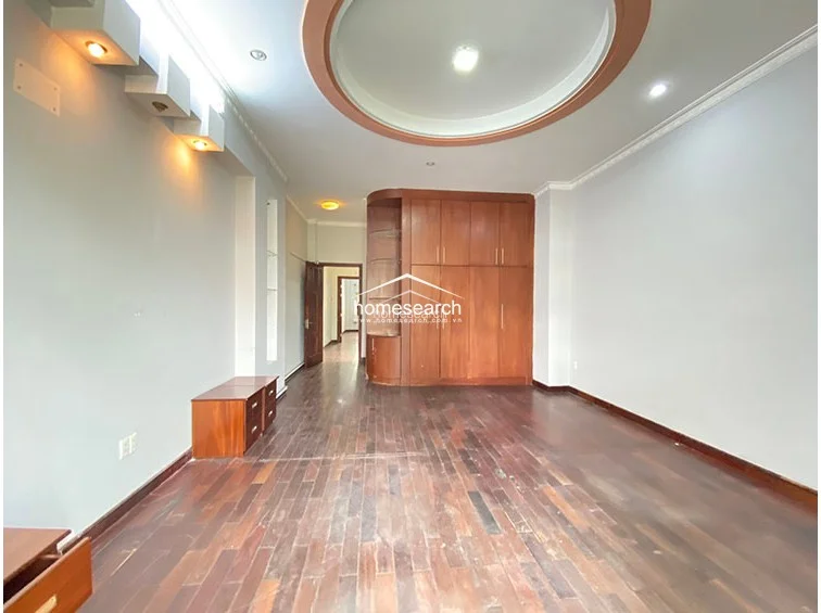 This bedroom features dark wood-look flooring and a prominent circular recessed ceiling design with integrated lighting. A large, floor-to-ceiling wooden wardrobe with integrated shelving provides substantial storage, while the room flows directly into a hallway leading to other areas of the home.