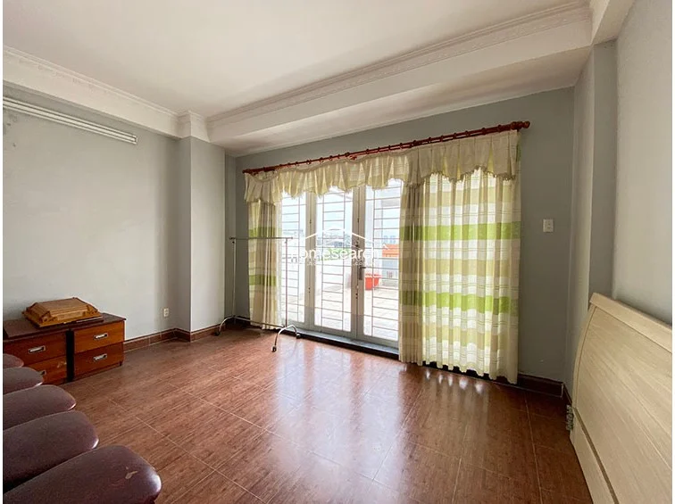 This room functions as a flexible living or bedroom space, featuring wood-look tile flooring and a large glass door that opens onto a private terrace. The walls are painted a soft grey, and the room is accented with traditional window treatments and a simple wooden bed frame.