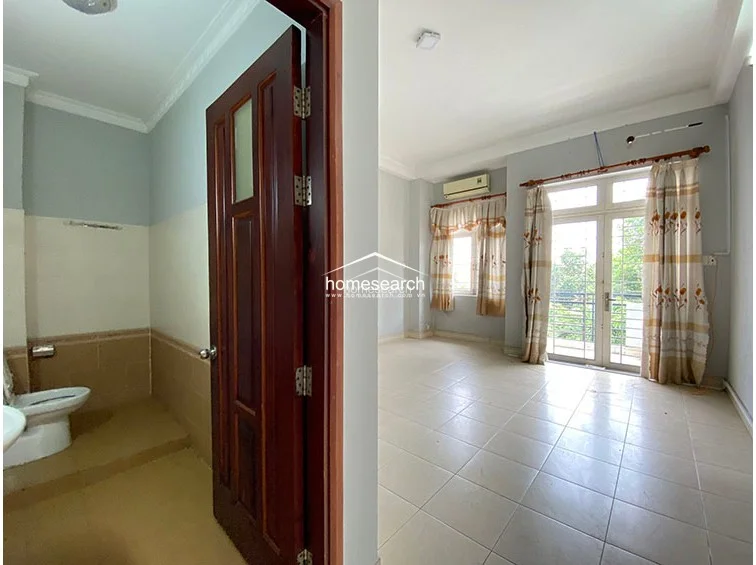 This split-view image shows a functional bathroom with tan-colored tiling and a separate bedroom area featuring light-colored floor tiles. The bedroom is equipped with an AC unit and a glass door leading to a balcony, offering a bright and practical layout for daily living.