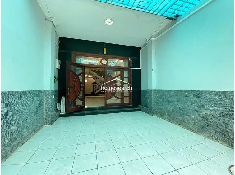 This image shows a covered outdoor entryway or garage area featuring light-colored floor tiles and walls partially clad in textured dark stone. The space is protected by a semi-transparent blue corrugated roof and connects directly to the interior living space through a large, decorative wooden glass-paneled door.