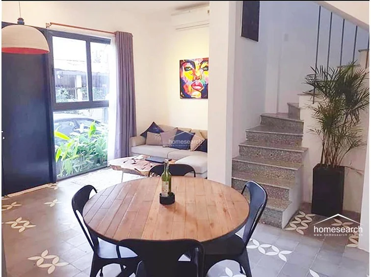 The living room is anchored by a comfortable sofa and a unique natural wood coffee table, positioned near a large window that invites ample natural light. A staircase with stone steps leads to the upper level, while a vibrant piece of wall art adds a modern, artistic touch to the white interior.