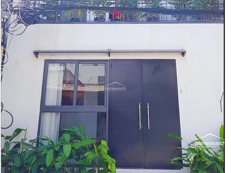 The exterior facade features a modern, minimalist white finish with a dark-framed glass door and window combination. A small metal awning provides shade, while the ground-level area is framed by lush green plants, creating a welcoming and private entrance.