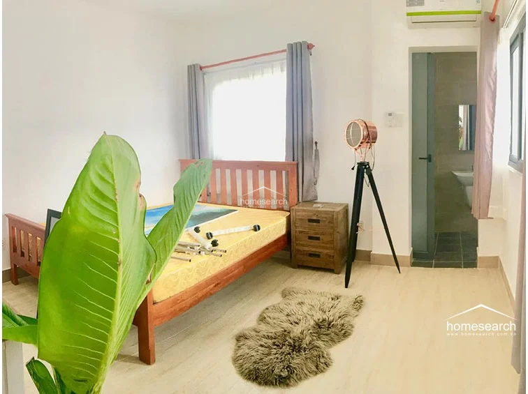 This bedroom features light wood-look flooring and a warm, wooden bed frame, creating a serene and inviting retreat. The room includes an en-suite bathroom, a decorative tripod floor lamp, and a large window with soft curtains that allow for plenty of natural light.