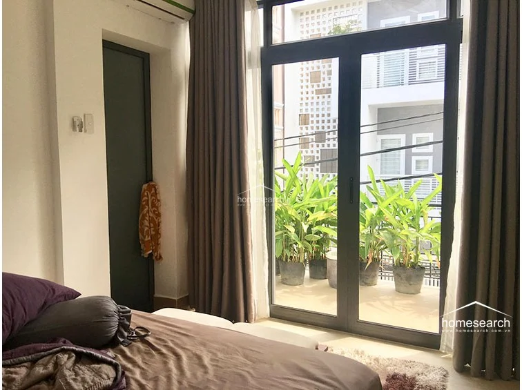 This bedroom features a floor-to-ceiling glass door that provides abundant natural light and direct access to a private balcony filled with potted greenery. The interior is finished with neutral walls, dark curtains, and a wall-mounted air conditioning unit for climate control.