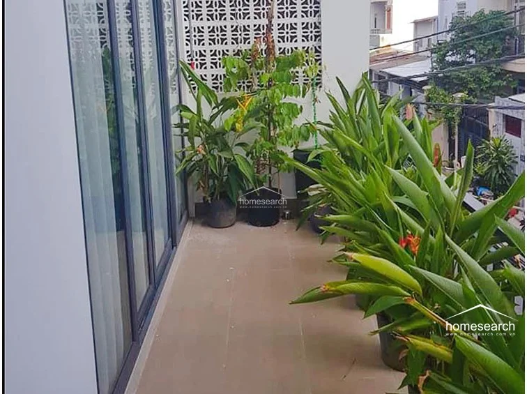 The private balcony is floored with neutral tiles and serves as a lush, semi-private outdoor retreat surrounded by potted plants. It is enclosed by a modern dark-framed glass door system that seamlessly connects the indoor living space to the exterior.