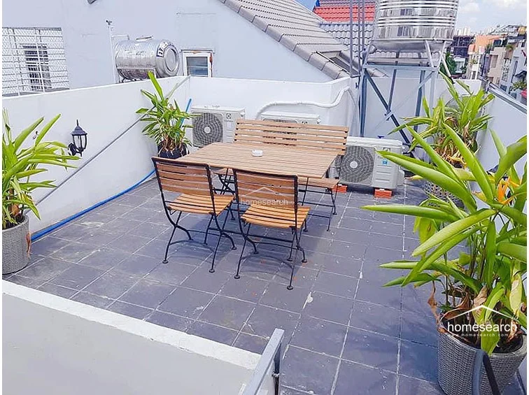 This rooftop terrace is an open-air space featuring dark tiled flooring and a wooden dining set, ideal for outdoor entertaining. The area is functional, equipped with multiple external air conditioning units and a large water tank, though it maintains a utilitarian aesthetic.