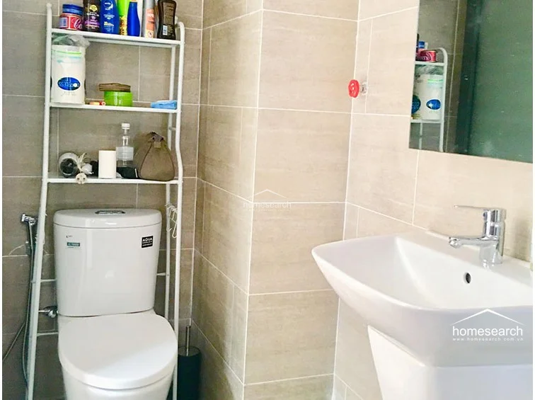 The bathroom is finished with modern, light-toned wall tiles and features a clean, white pedestal sink and a standard toilet. A space-saving metal shelving unit is positioned over the toilet to provide essential storage in this compact, well-maintained space.
