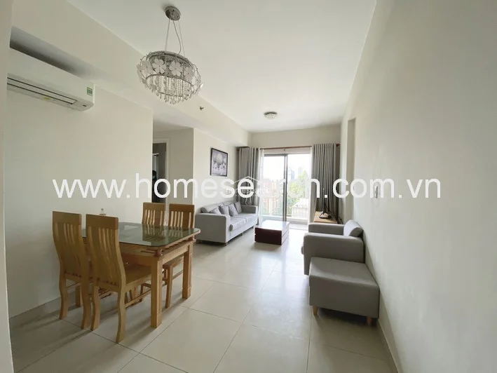 This open-concept living and dining area features neutral off-white walls and light-colored tile flooring, creating a bright and airy atmosphere. The space is furnished with a light grey sofa set, a wooden dining table, and a decorative crystal chandelier, with a wall-mounted AC unit and a sliding glass door providing access to an outdoor balcony.