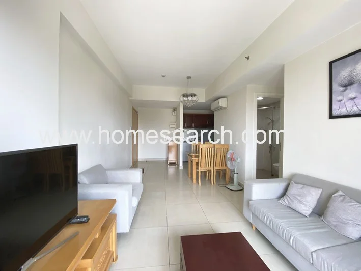 This alternate angle of the living room highlights the flow into the adjacent kitchen and hallway, maintaining the consistent neutral color palette and tile flooring. The room is equipped with a wall-mounted television, a wooden media console, and a ceiling-mounted AC unit, ensuring a functional and comfortable living environment.