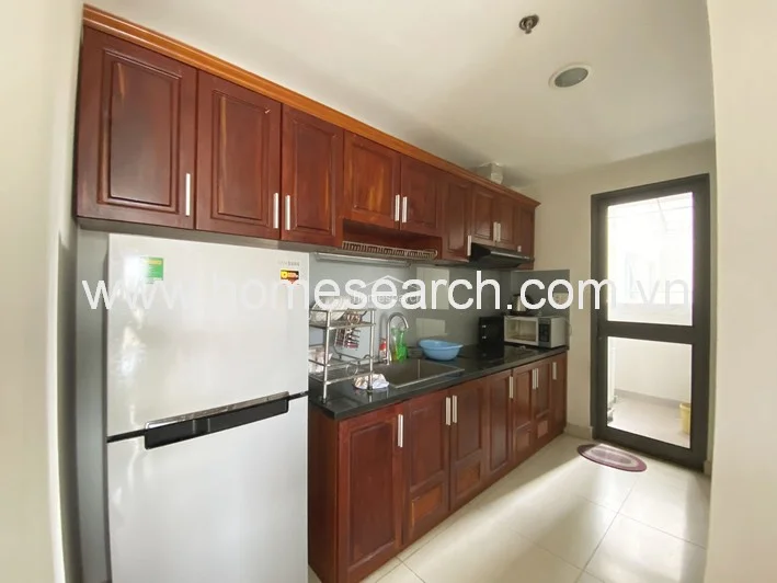 The kitchen features a practical galley layout with dark-stained wooden cabinetry providing ample storage, complemented by a black countertop and a stainless steel sink. It is equipped with a full-sized refrigerator and a built-in stovetop, with a glass door leading to a small utility balcony for additional ventilation.