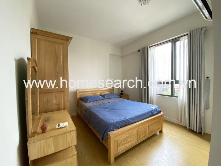 This bedroom features warm wood-look laminate flooring and is furnished with a sturdy wooden bed frame, a matching wardrobe, and a vanity set. Large windows with floor-to-ceiling curtains allow for significant natural light, while a wall-mounted AC unit ensures climate control.