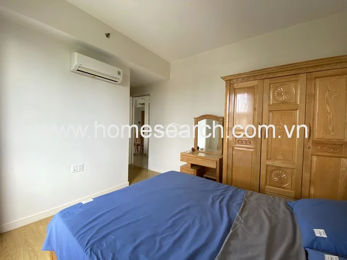 This view of the bedroom emphasizes the solid wood furniture, including a large multi-door wardrobe and a vanity with a mirror, which offer substantial storage. The room maintains a clean, minimalist aesthetic with white walls and a consistent light wood flooring finish, presenting a well-maintained and tidy space.